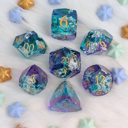 A set of iridescent polyhedral dice arranged on a white faux-fur surface with pastel star-shaped props. The dice have shifting teal, blue, purple, and silver foil effects with gold-colored numbering.