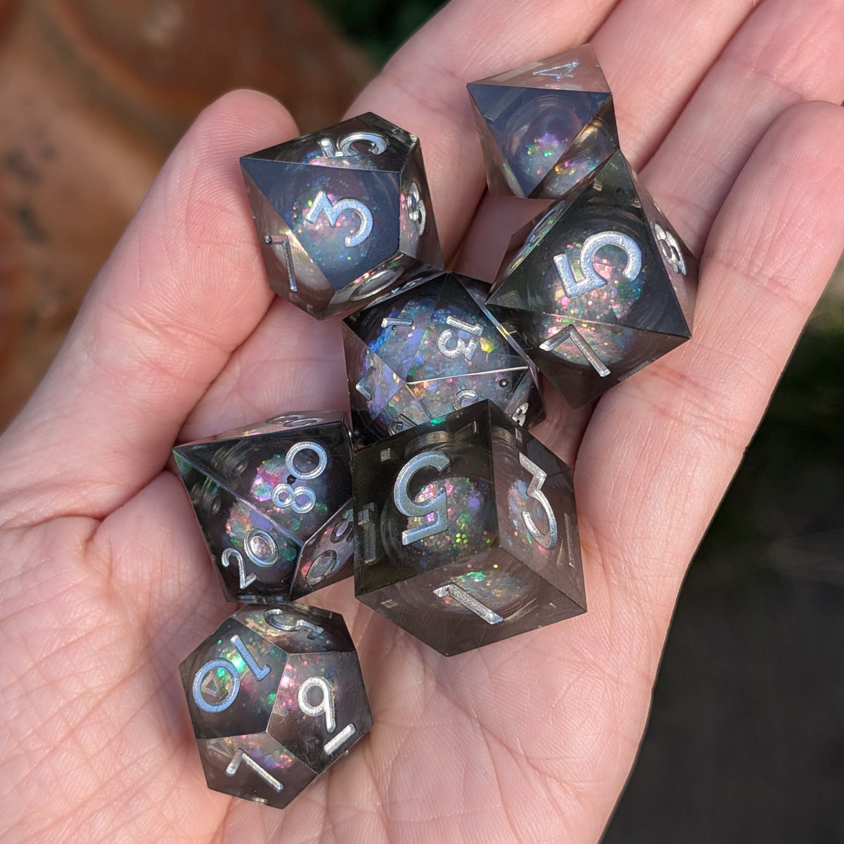 A set of seven translucent dark gray polyhedral gaming dice with holographic glitter and white numbering, held in a person's hand.