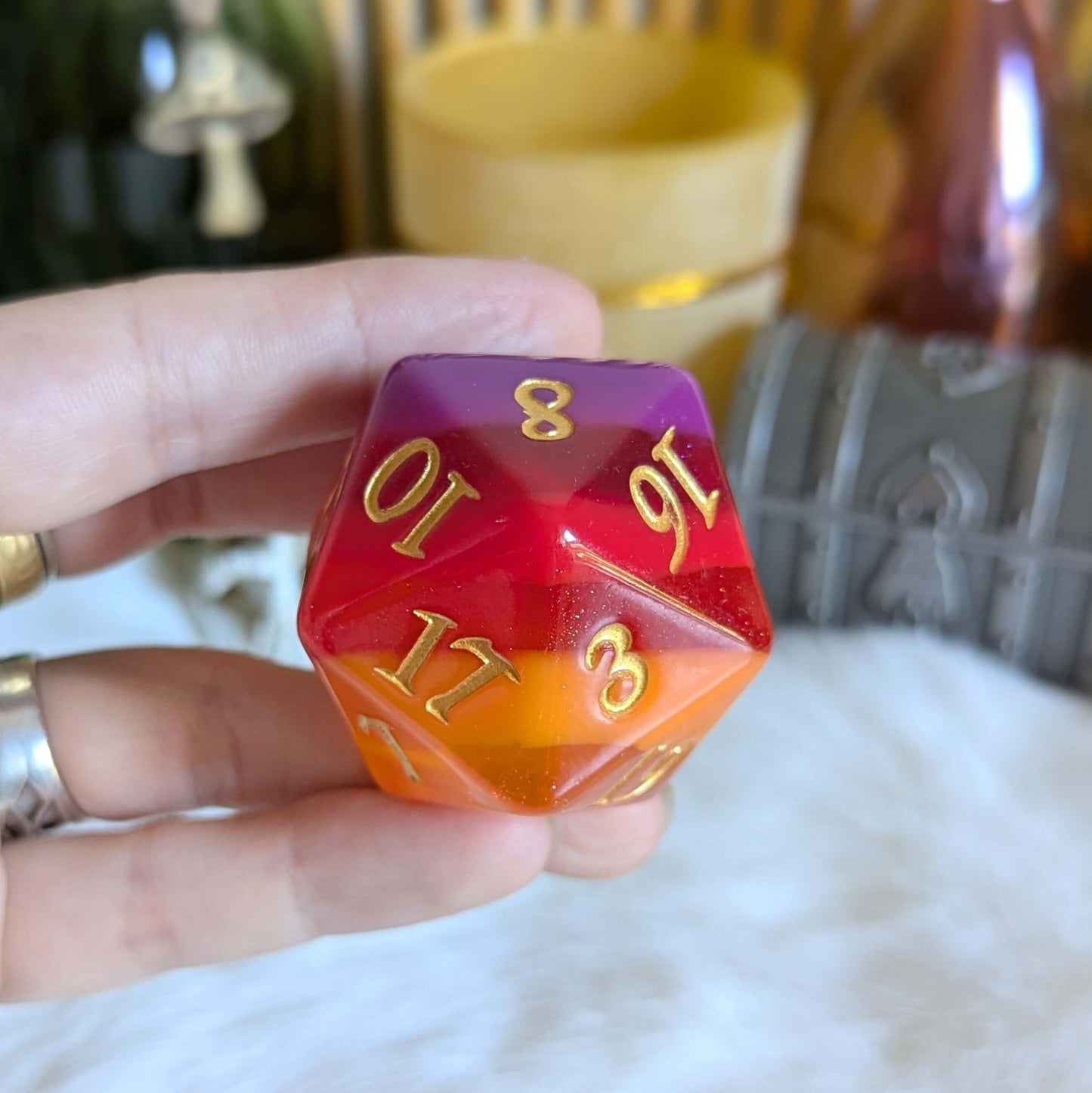 Emberstorm Large D20 die with gold fantasy font, featuring a vibrant flow of deep purple, red, and orange hues, held in a hand.