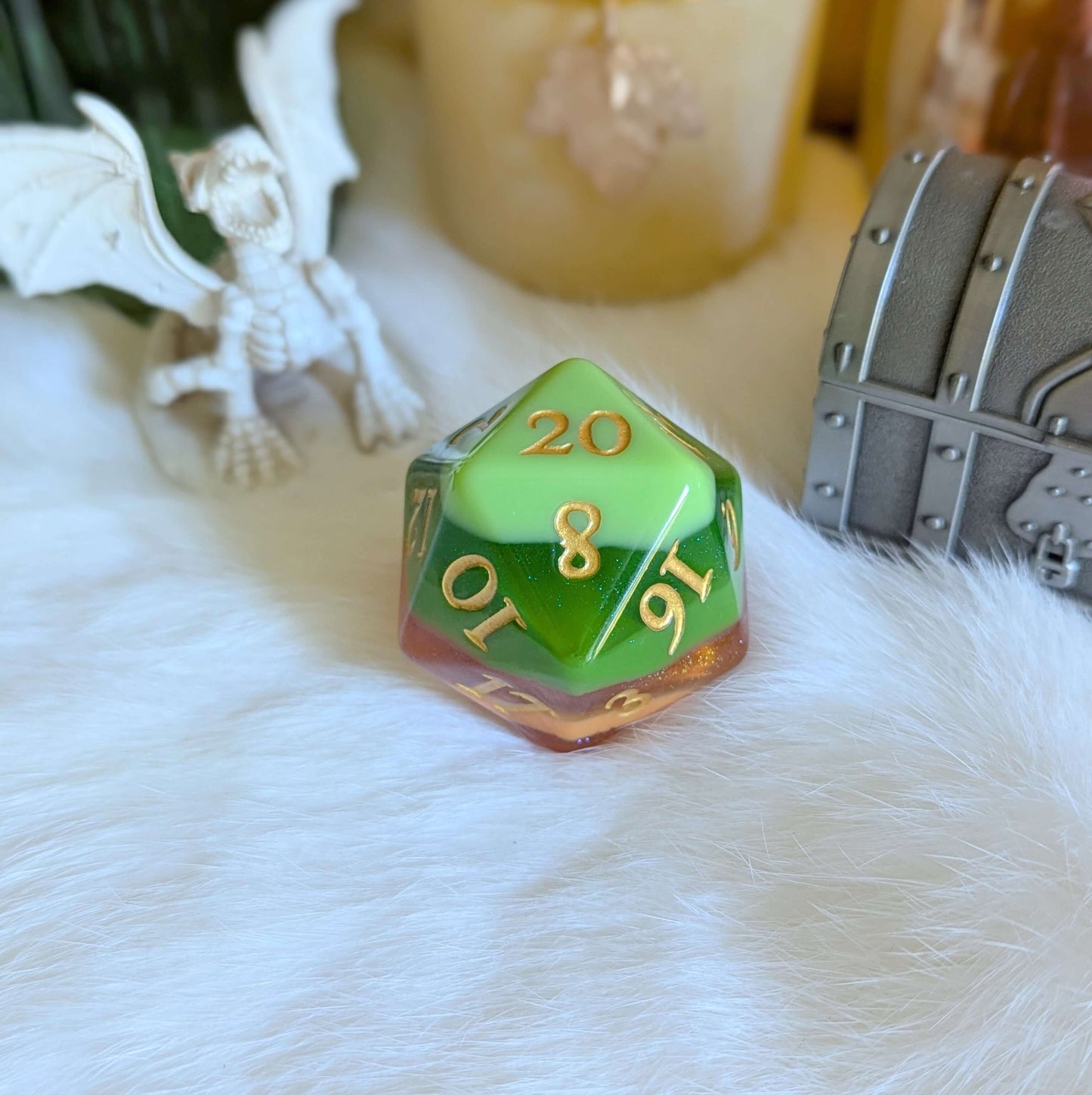 Green and gold D20 die on a fluffy white surface with dragon and treasure chest figures in the background.