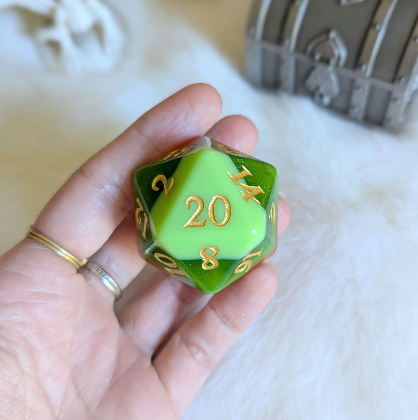 Hand holding green and yellow D20 die with gold numbers, displayed on a white fluffy background with a treasure chest in the corner.