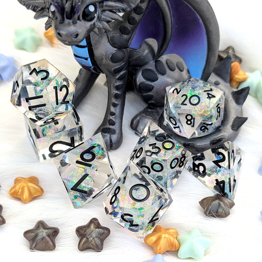 A set of holographic polyhedral dice with black printed numbers, arranged on a white textured surface with a dark dragon figurine and small star-shaped accessories.