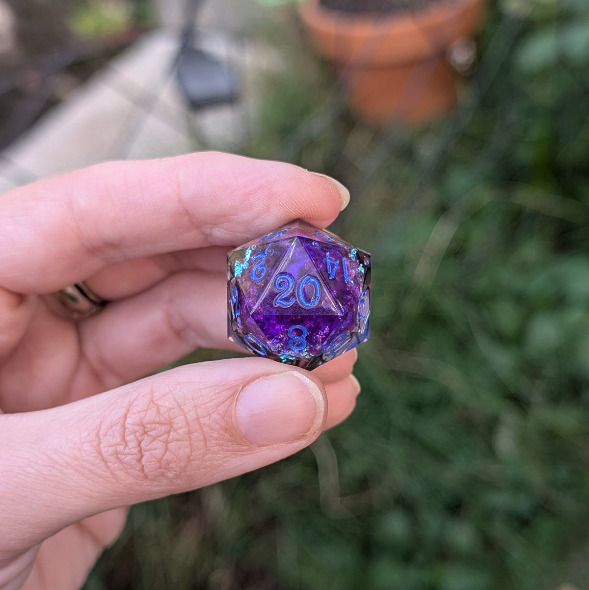 A hand holding a translucent purple and blue 20-sided d20 die outdoors with a blurred garden background.