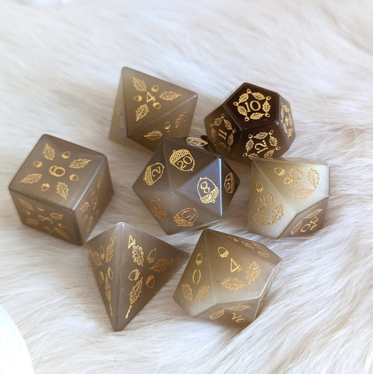 Light brown cat's eye dice set with acorn and oak leaf carvings, featuring d20, d12, d10, d%, d8, d6, and d4 on a soft surface.