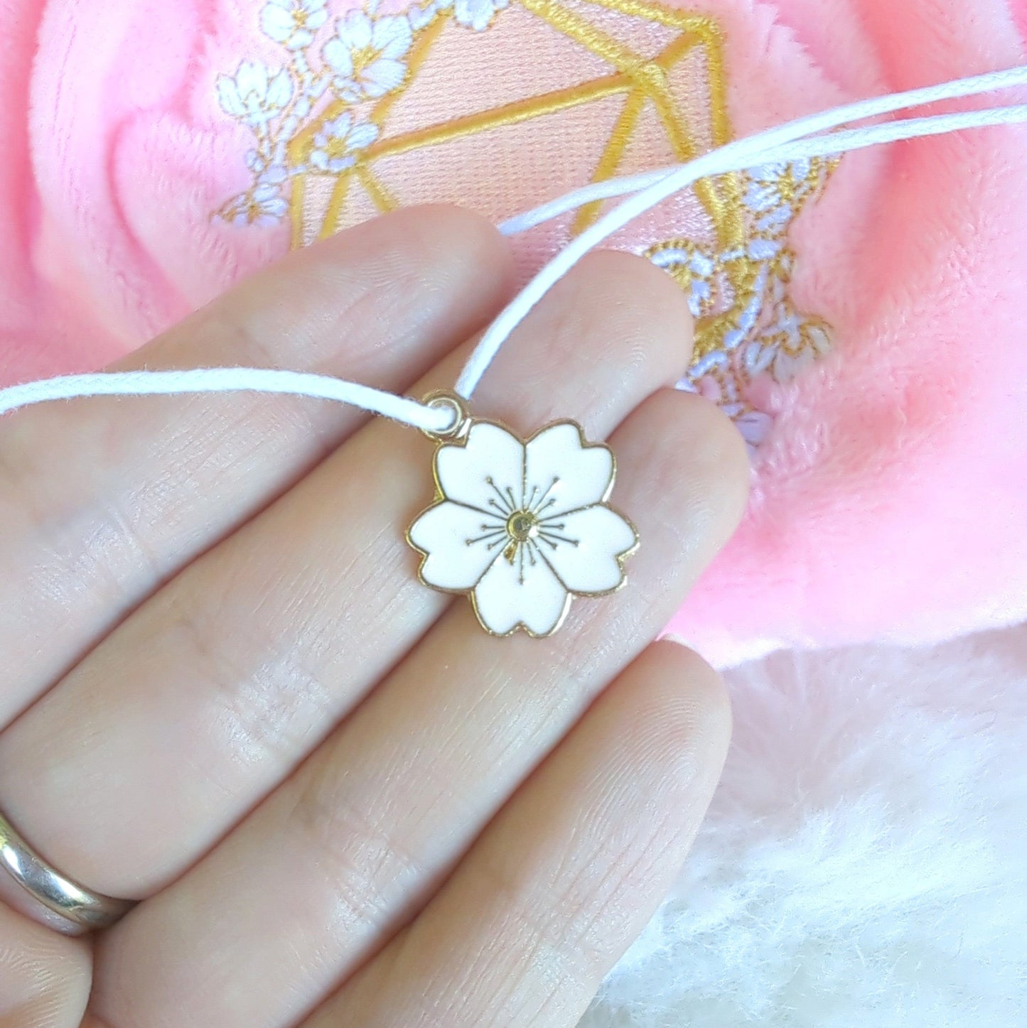 Hand holding cherry blossom charm with pink plush fabric and embroidered D20 design in the background.