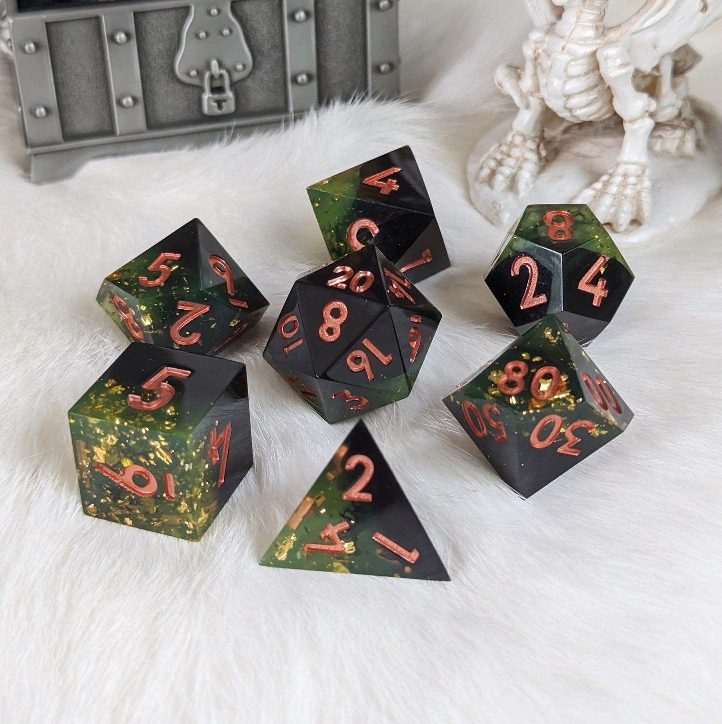 Dark green and black DnD dice set with gold flakes and copper font, displayed on a white surface in front of a treasure box and figurine.