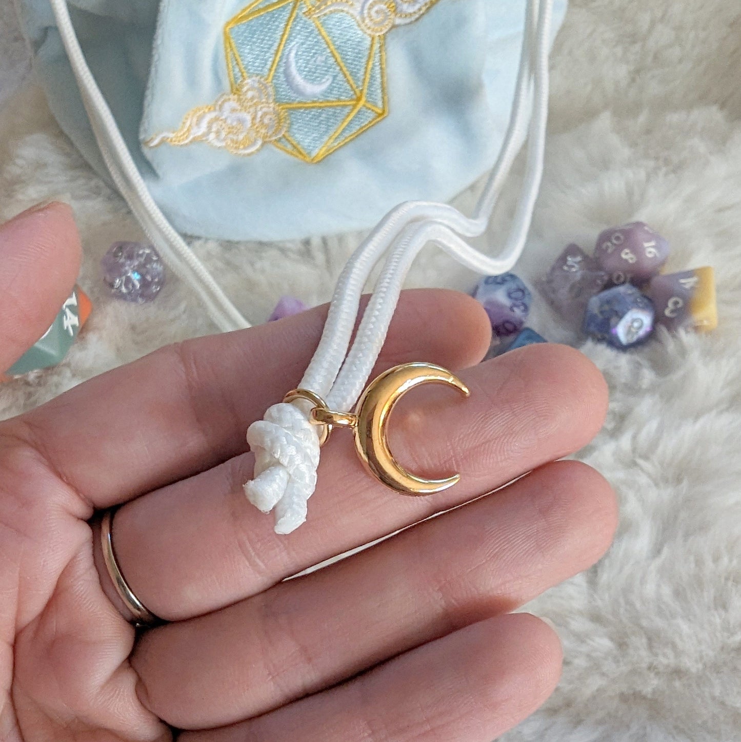 Close-up of gold crescent bead on drawstring of plushy blue dice bag with embroidered D20 and scattered polyhedral dice.