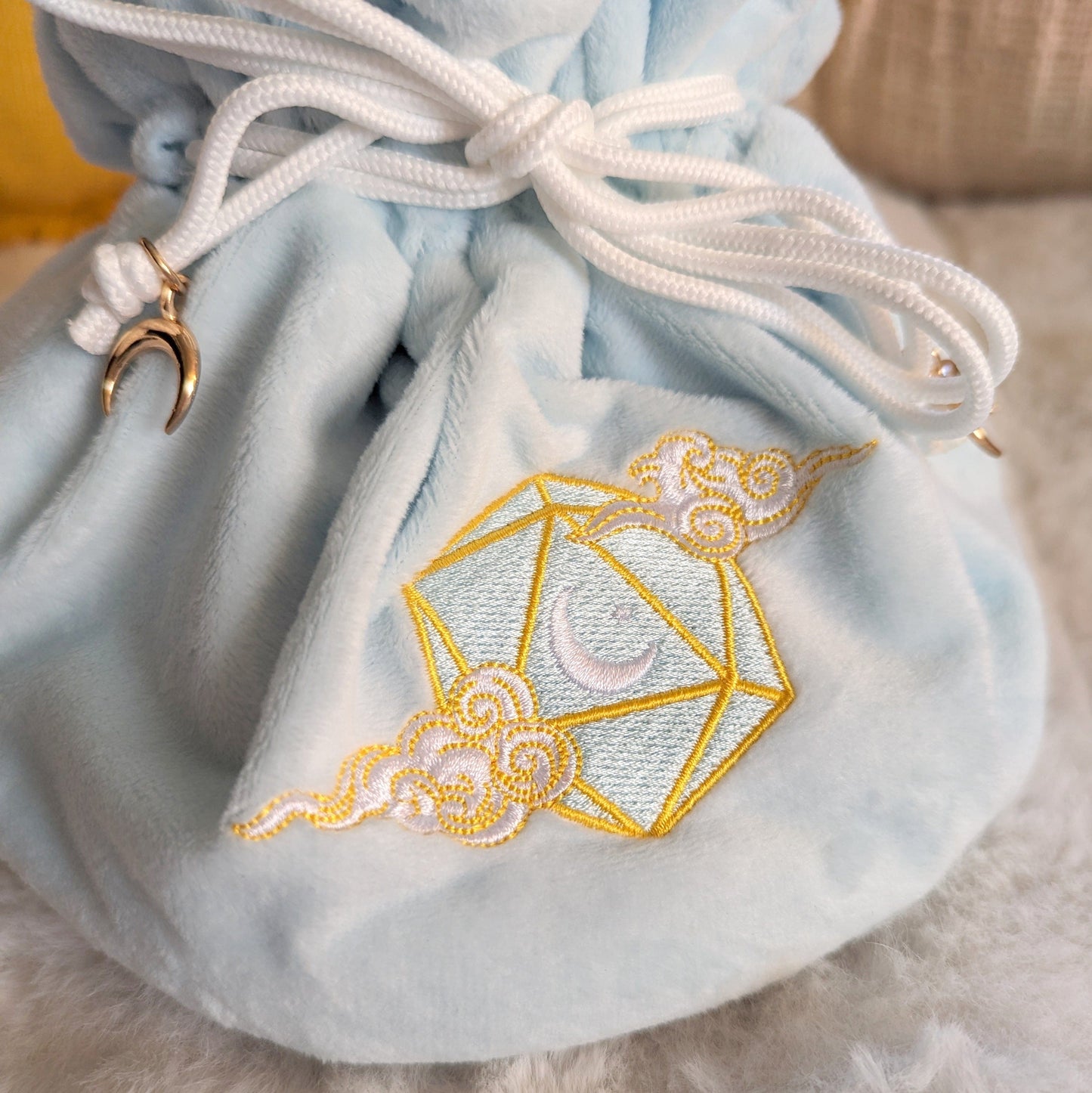 Blue plush dice bag with an embroidered D20, crescent moon, and cloud design, featuring white drawstrings and gold crescent beads.