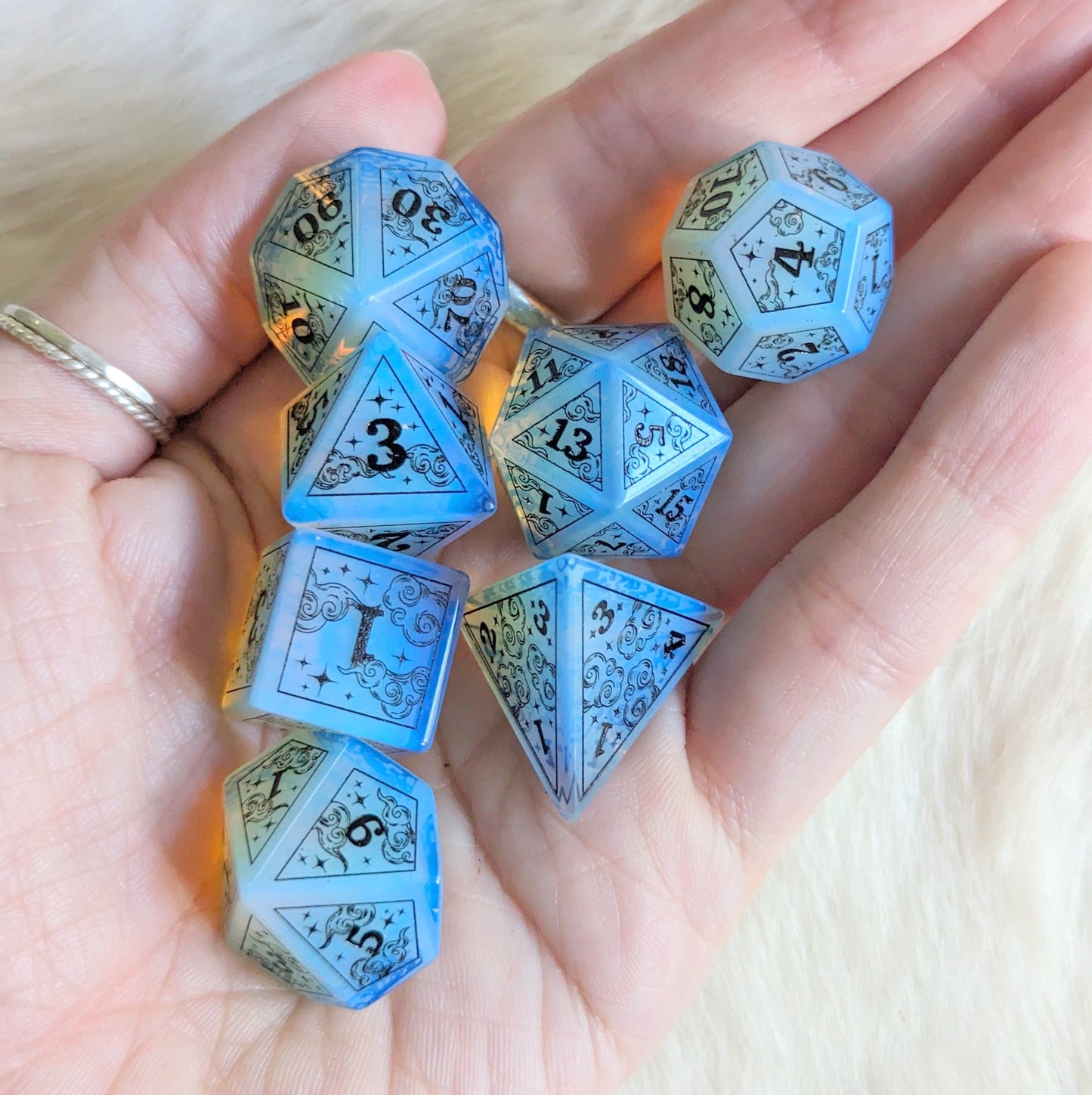 Hand holding Dreamy Opalite Dice Set with black font, featuring cloud and moon designs, perfect for Dungeons & Dragons.