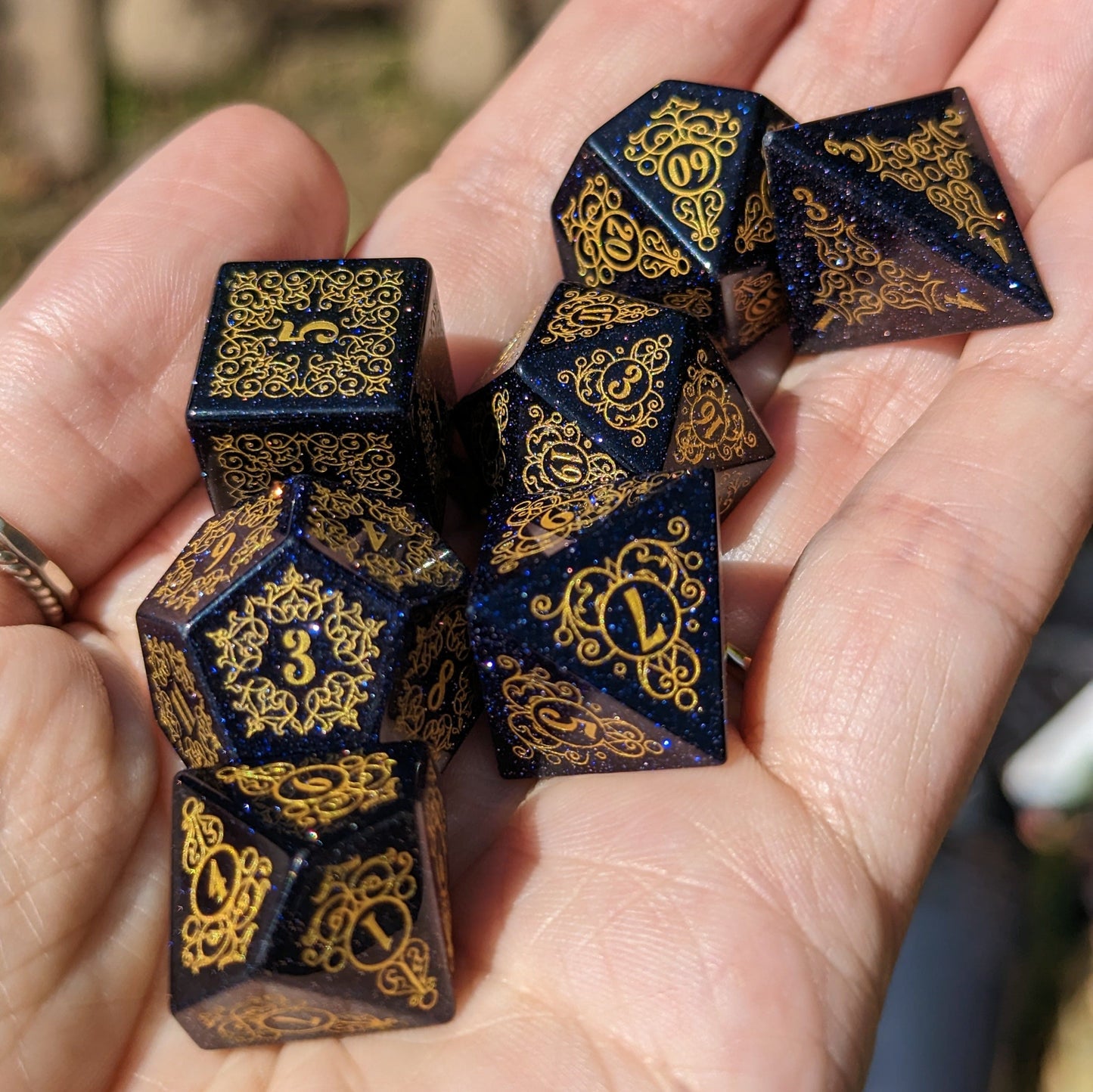 Hand holding Garden Gate Blue Sandstone Dice Set with intricate gold designs and dark blue shimmering surface.