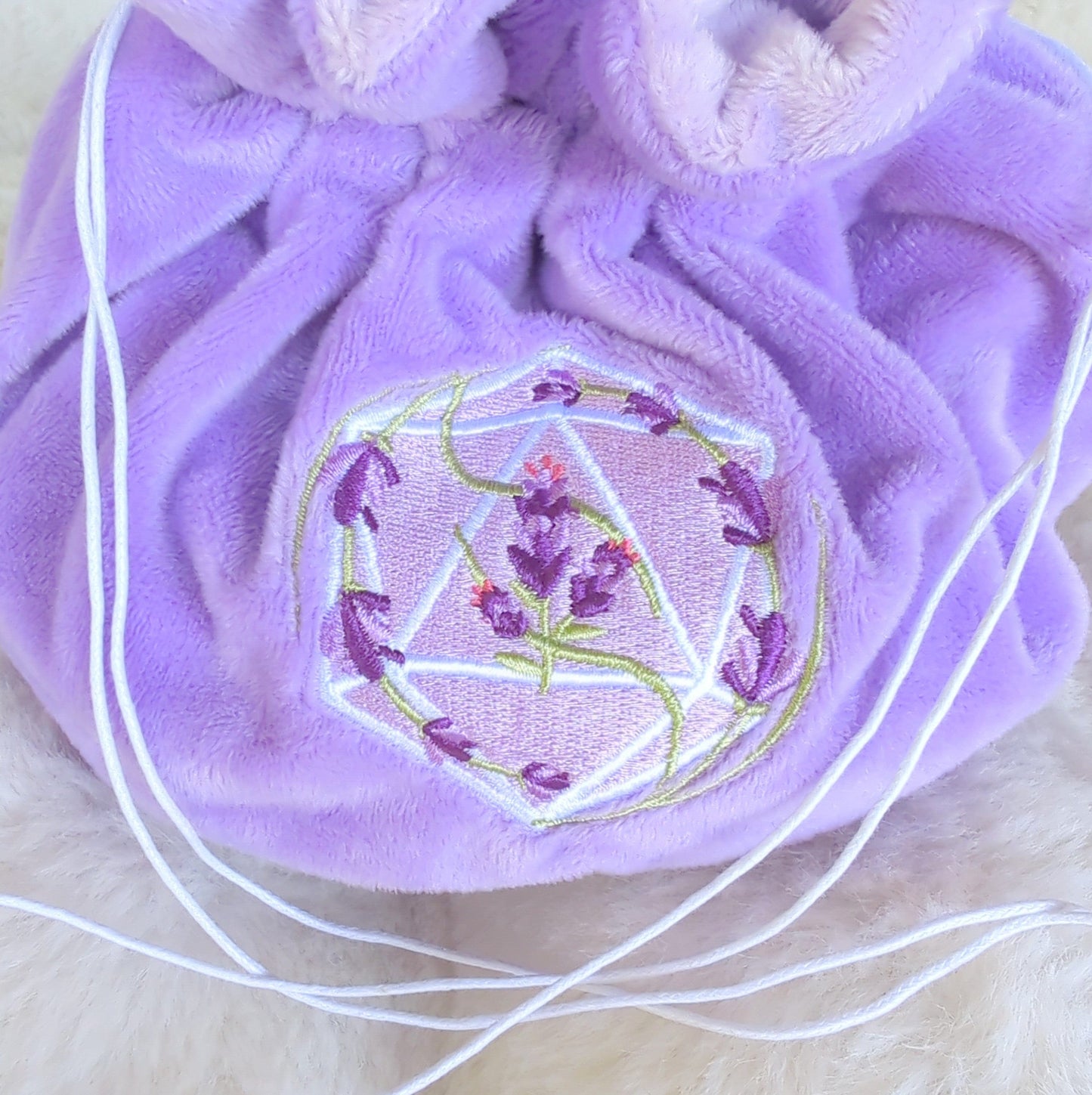 Lavender plush dice bag with D20 and swirling lavender embroidery, featuring white drawstrings and butterfly charms.