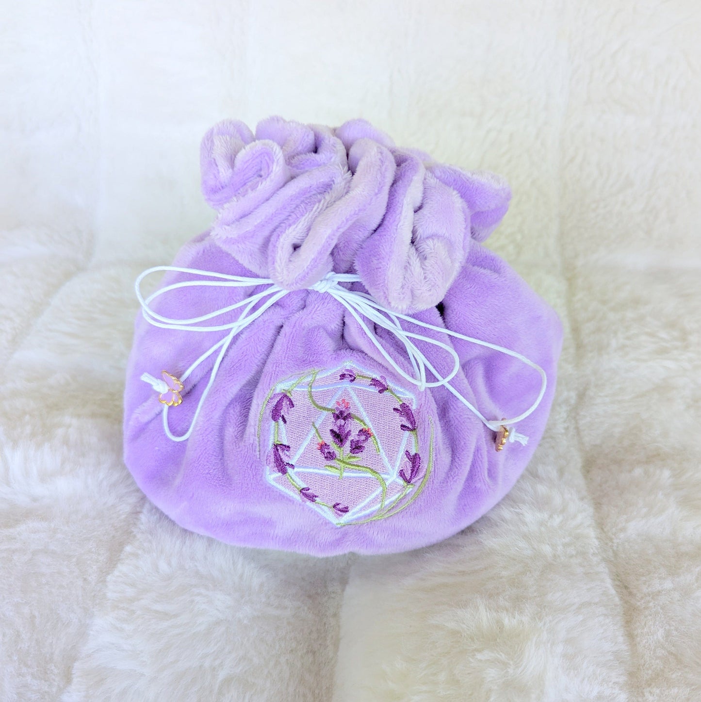 Lavender dice bag with D20 embroidery and purple butterfly charms, plush fabric, and drawstrings on a soft surface.
