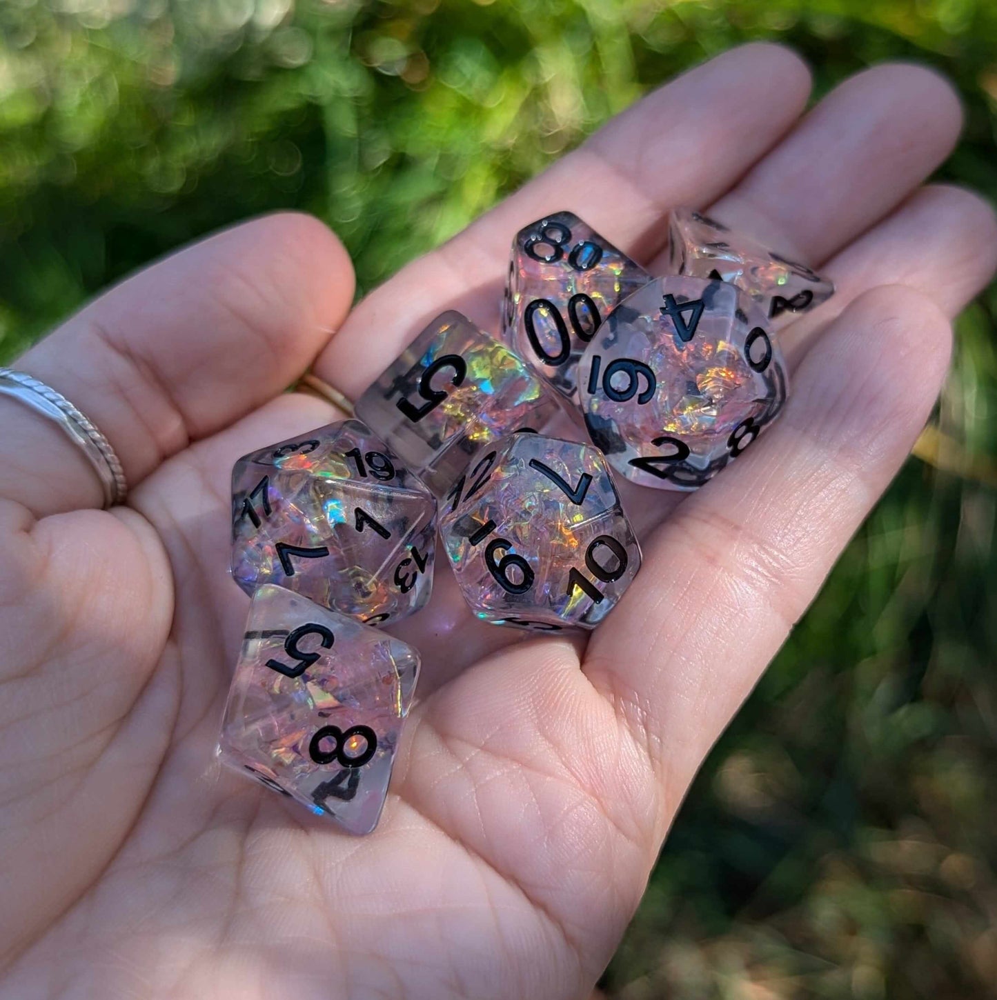 Hand holding Moonlight Fantasy TTRPG Dice Set with pink iridescent cellophane and black font against green background.