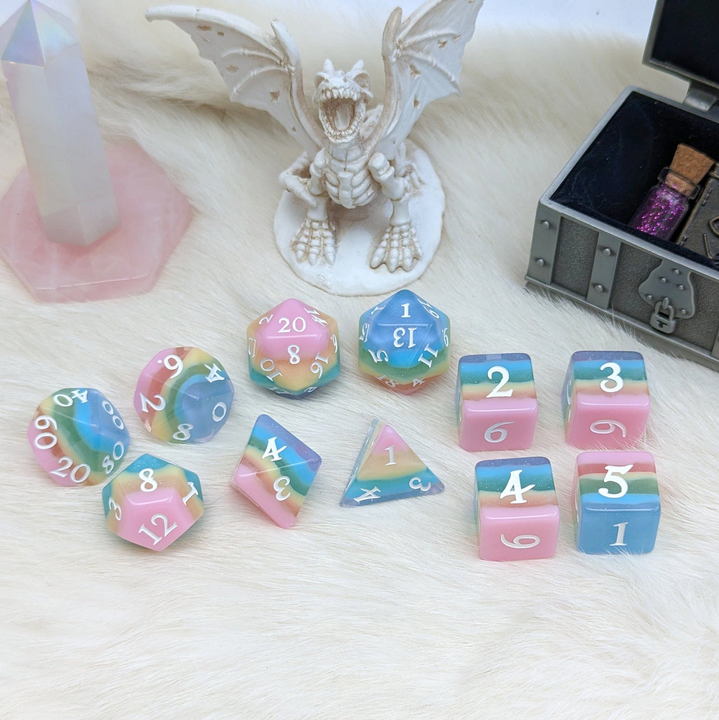 Pastel rainbow striped DND dice set with opaque and clear glittering layers, including 11 pieces on a fluffy surface with fantasy decor.