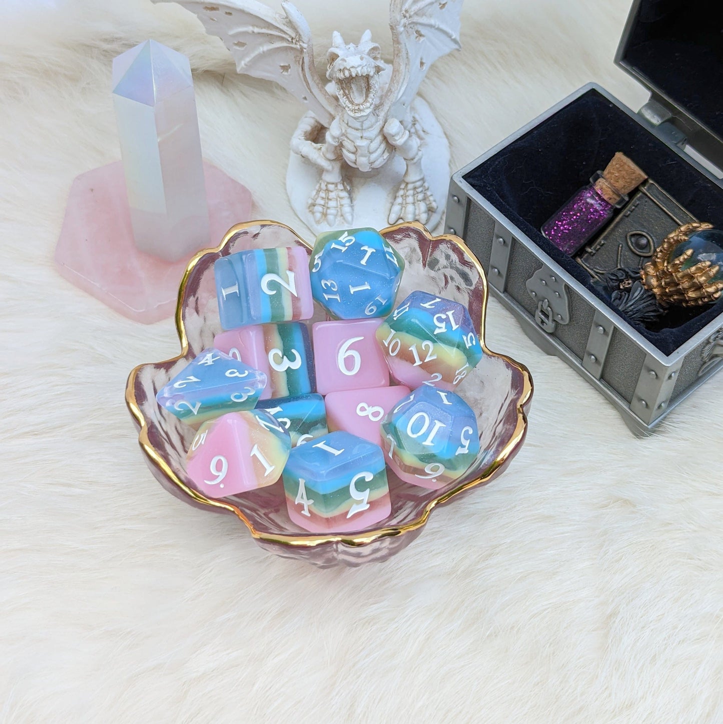 Pastel rainbow stripe DnD dice set with glitter layers in glass dish, surrounded by fantasy-themed decor.