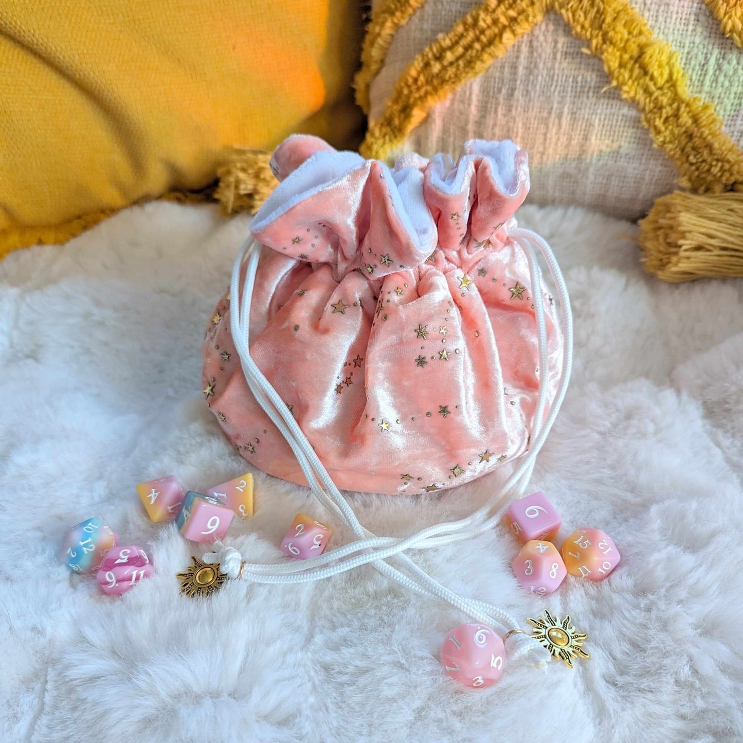 Starry pink dice bag with gold star design, white drawstrings, and solar charms on soft fabric, surrounded by colorful dice.