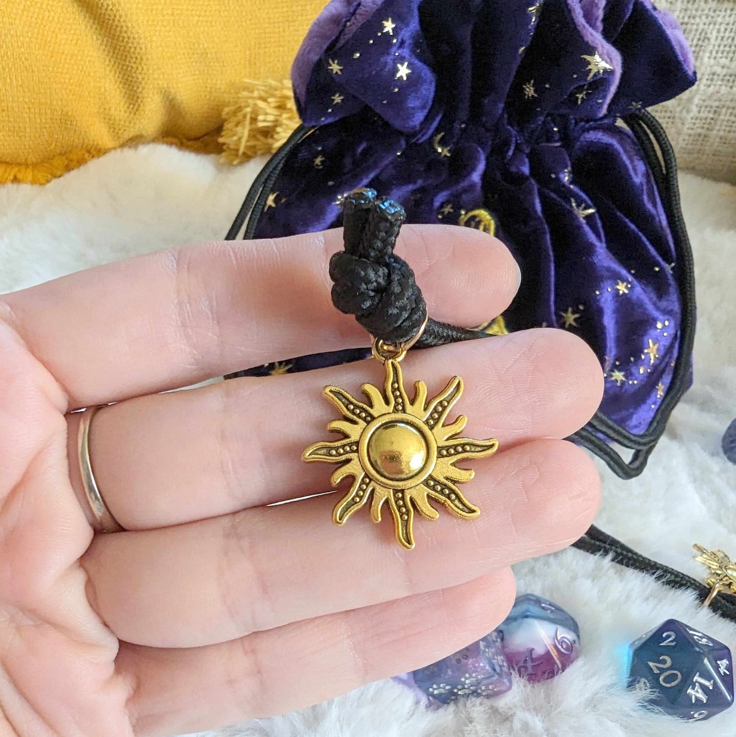 Hand holding golden sun charm on purple velvet wizard dice bag with starry design and D20 dice.