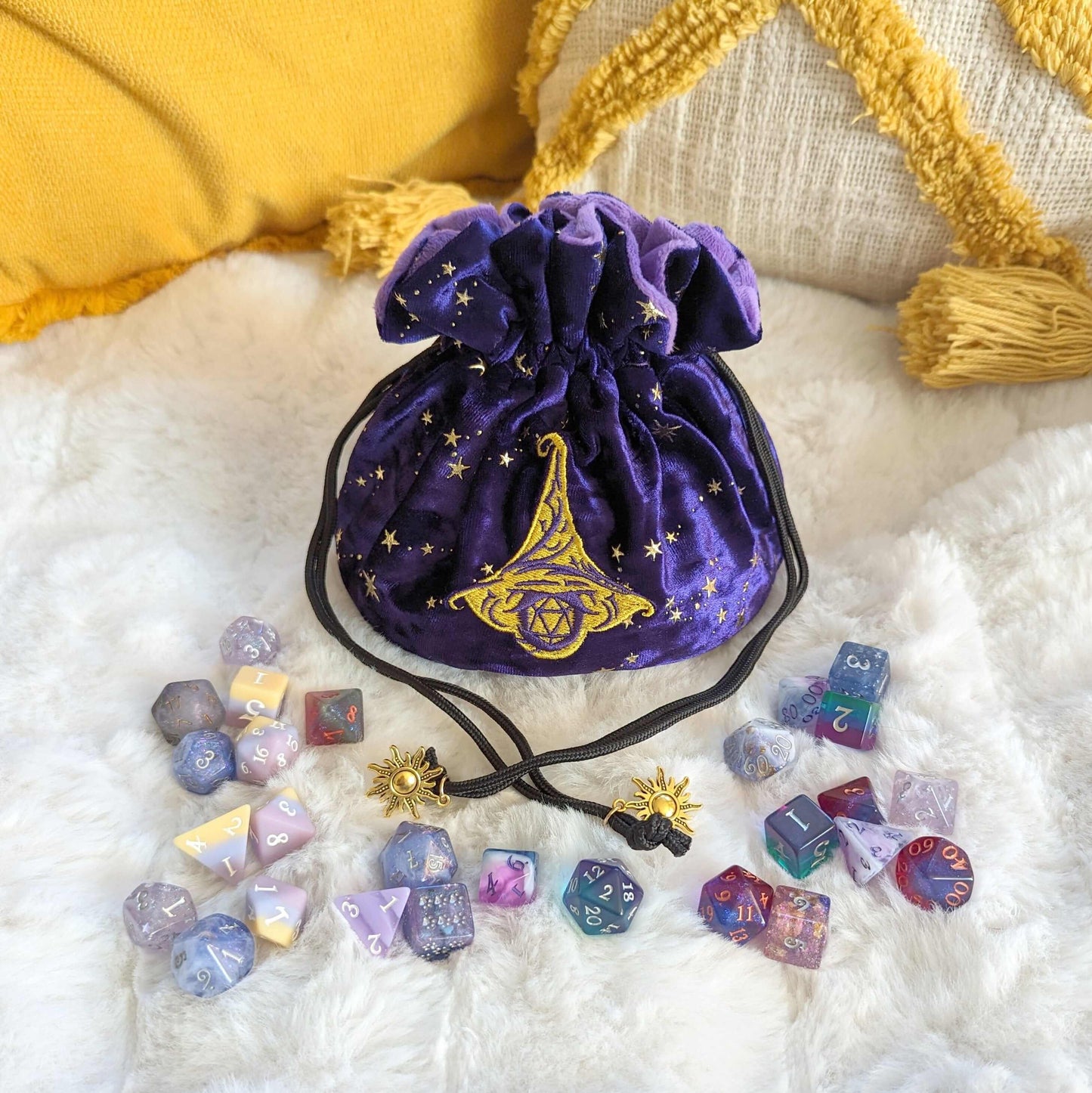Purple velvet wizard dice bag with embroidered hat and D20, surrounded by colorful dice, featuring golden sun charm drawstrings.