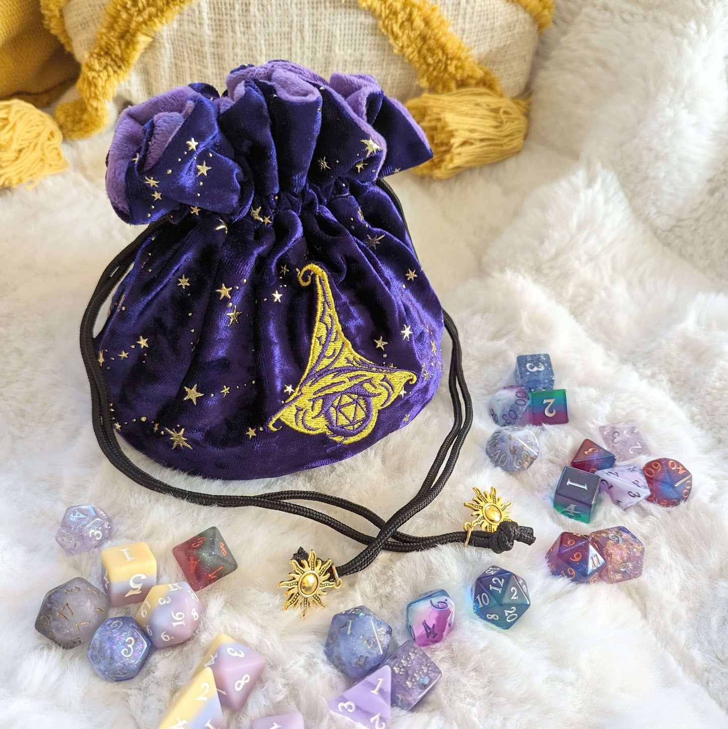 Purple velvet multi-pocket dice bag with embroidered wizard hat and D20, golden sun charms, surrounded by colorful dice.