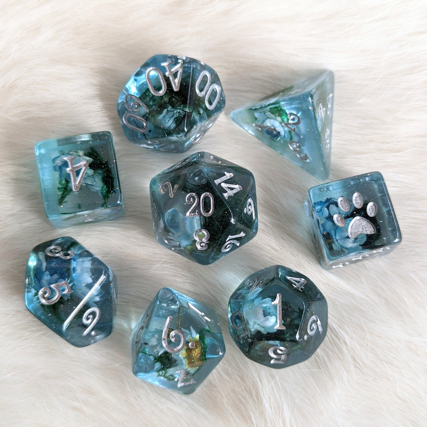 Blue flower and moss DND dice set with dried flowers, moss in clear blue resin, extra D6 with paw design, on a fuzzy background.