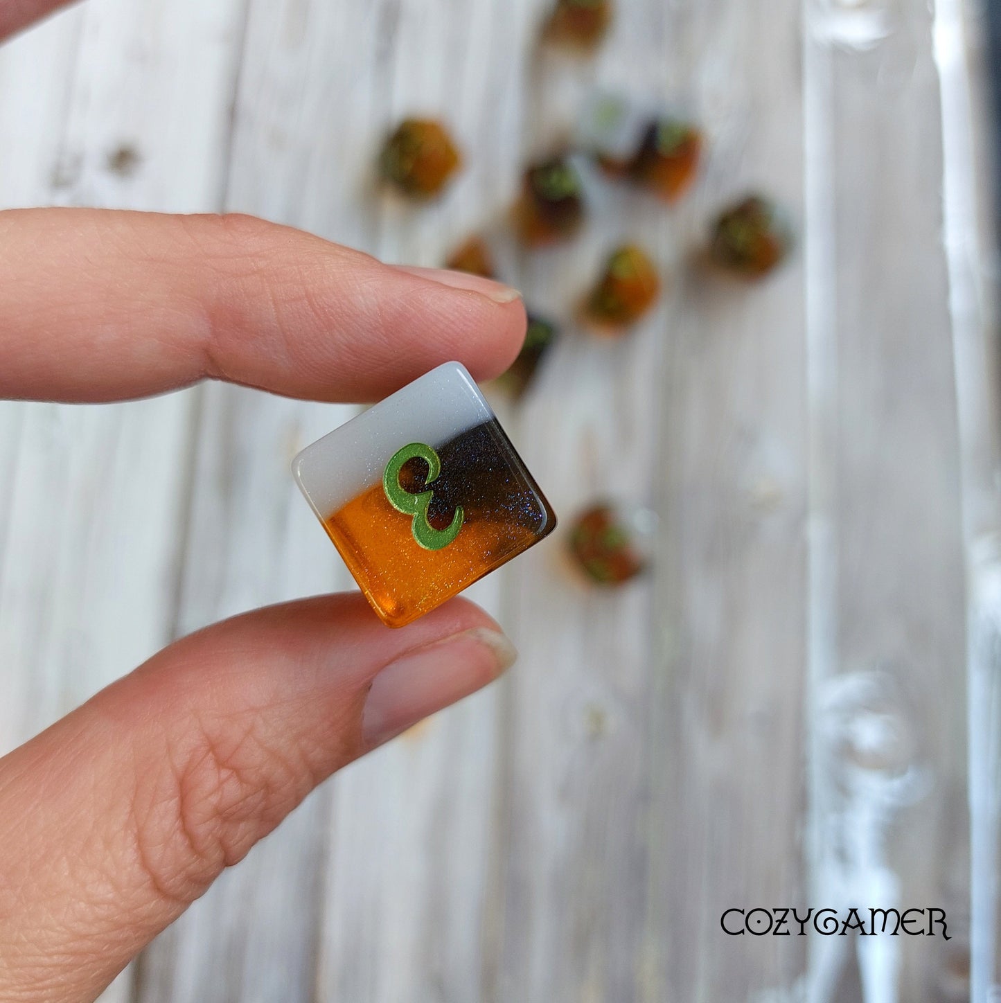 Close-up of a hand holding a beer-themed dice from the Calico Hazy IPA Dice Set with amber and white colors.