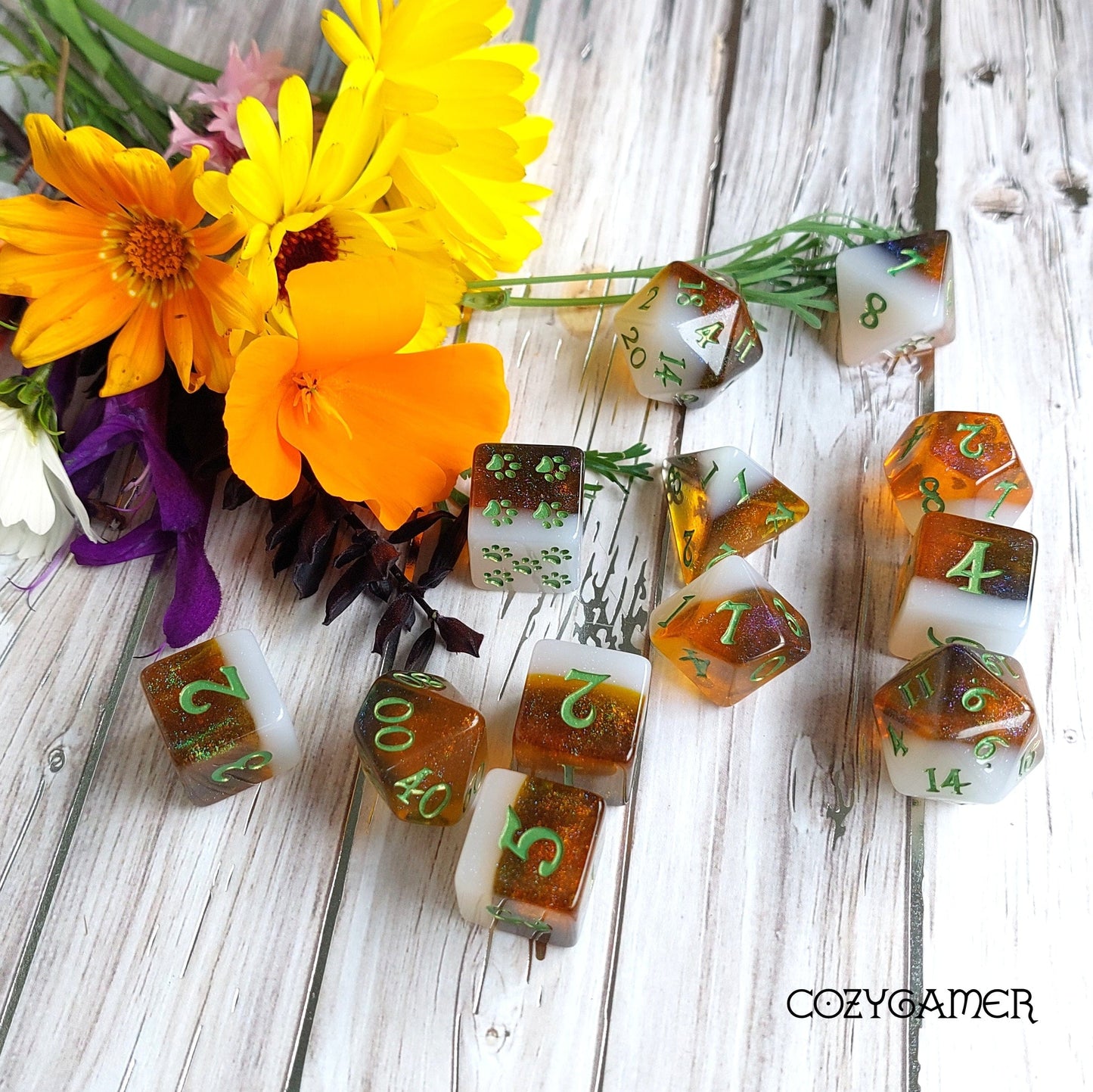 Calico Hazy IPA Dice Set with 12 beer-themed DND dice in calico cat colors, displayed with vibrant flowers on a wooden surface.
