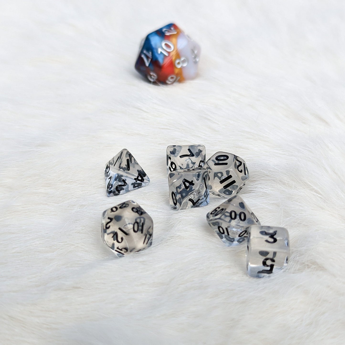 Clear mini polyhedral DND dice set with seven pieces on soft white background, showcasing cute and readable size compared to a regular D20.