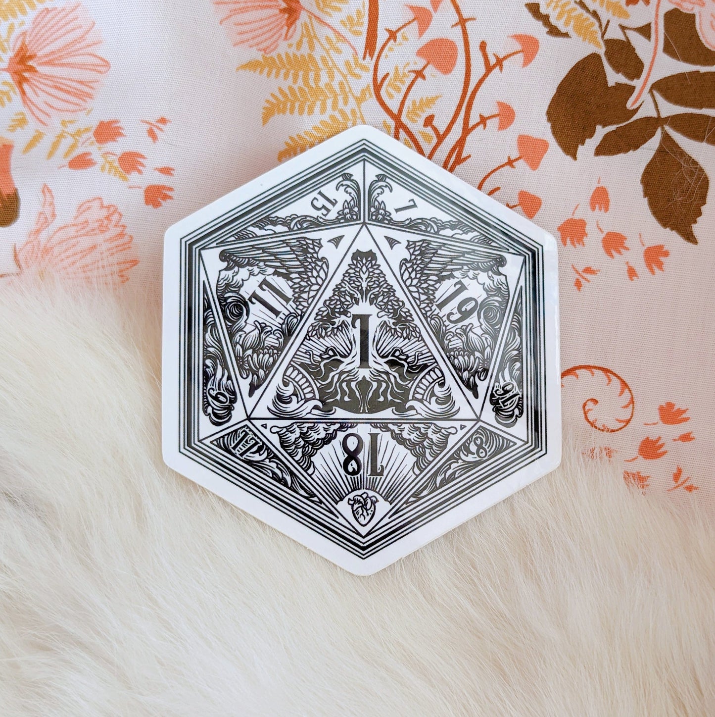 Intricate D20 dice sticker with detailed fantasy designs on floral background.
