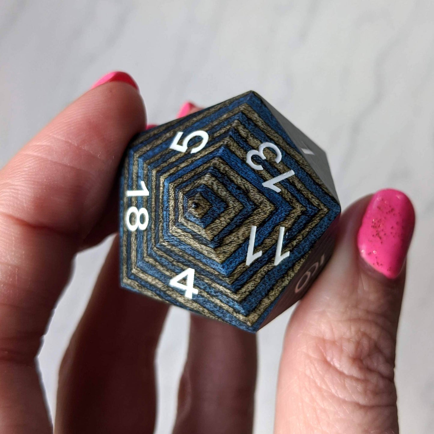 Hand holding dark blue and grey wood stripe large wooden D20 dice with visible numbers and intricate pattern.