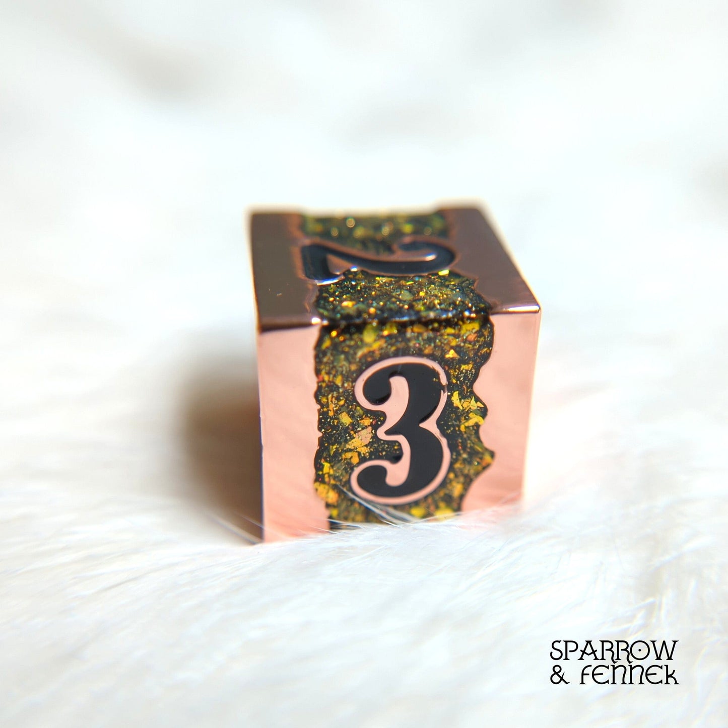 Copper and green/gold glitter polyhedral die on white fur background, part of Fissure Metal Dice Set for tabletop games.