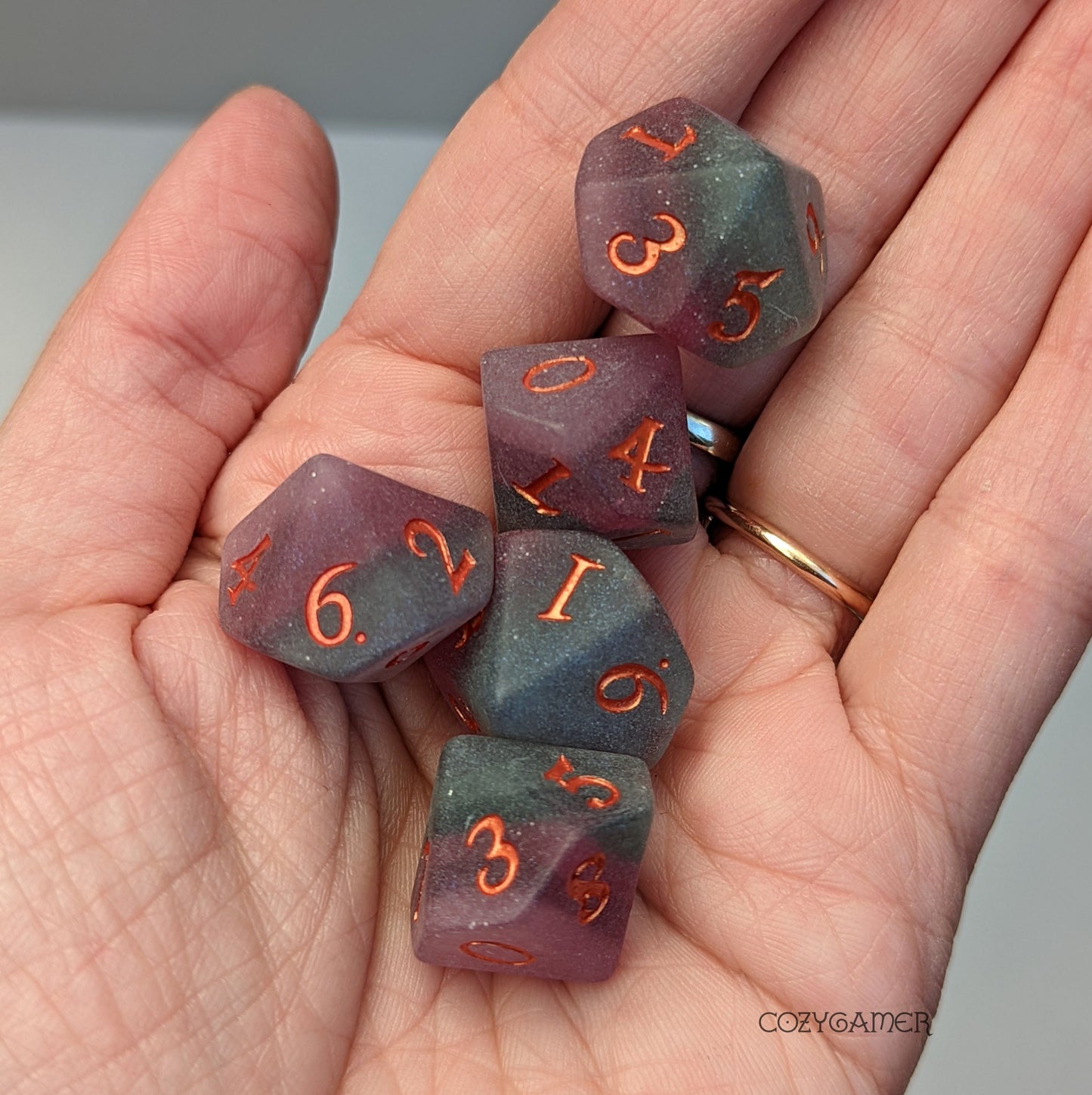 Hand holding five Fluorite D6 dice with purple and green matte finish and custom font numbers designed for Dungeons and Dragons.