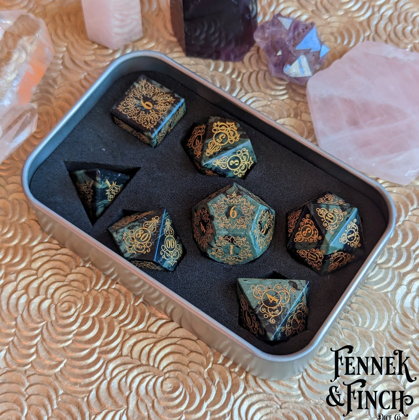 Green jasper gemstone dice set with intricate garden gate design in a metal box, surrounded by crystals.