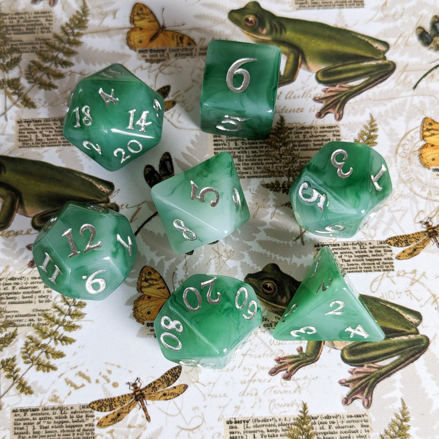 Jade 7-piece DnD dice set with green and white resin, silver fantasy numbers, displayed on a nature-themed background with frogs.