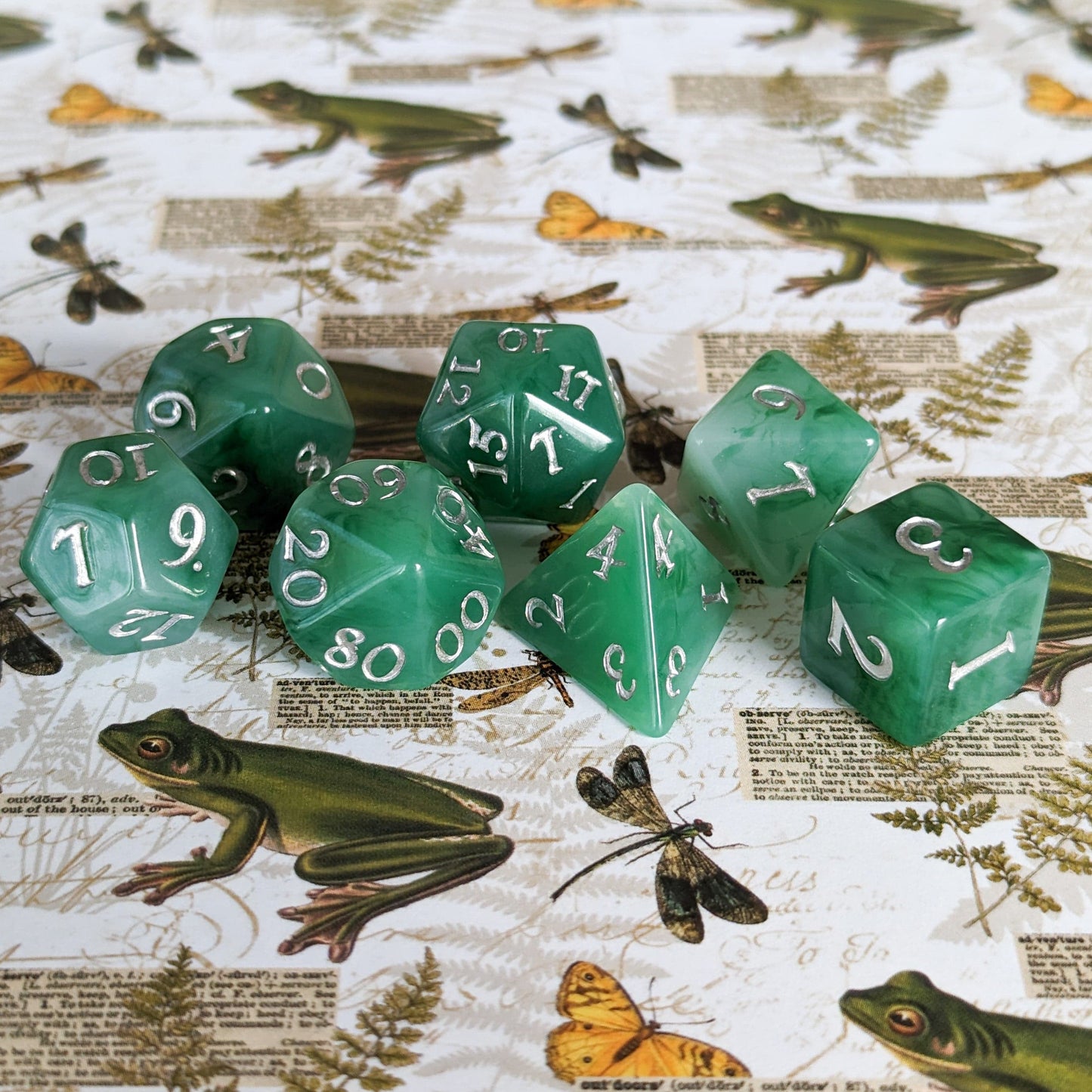 Jade 7-piece DnD dice set in green resin with silver numbers, displayed on a nature-themed background with frogs and butterflies.