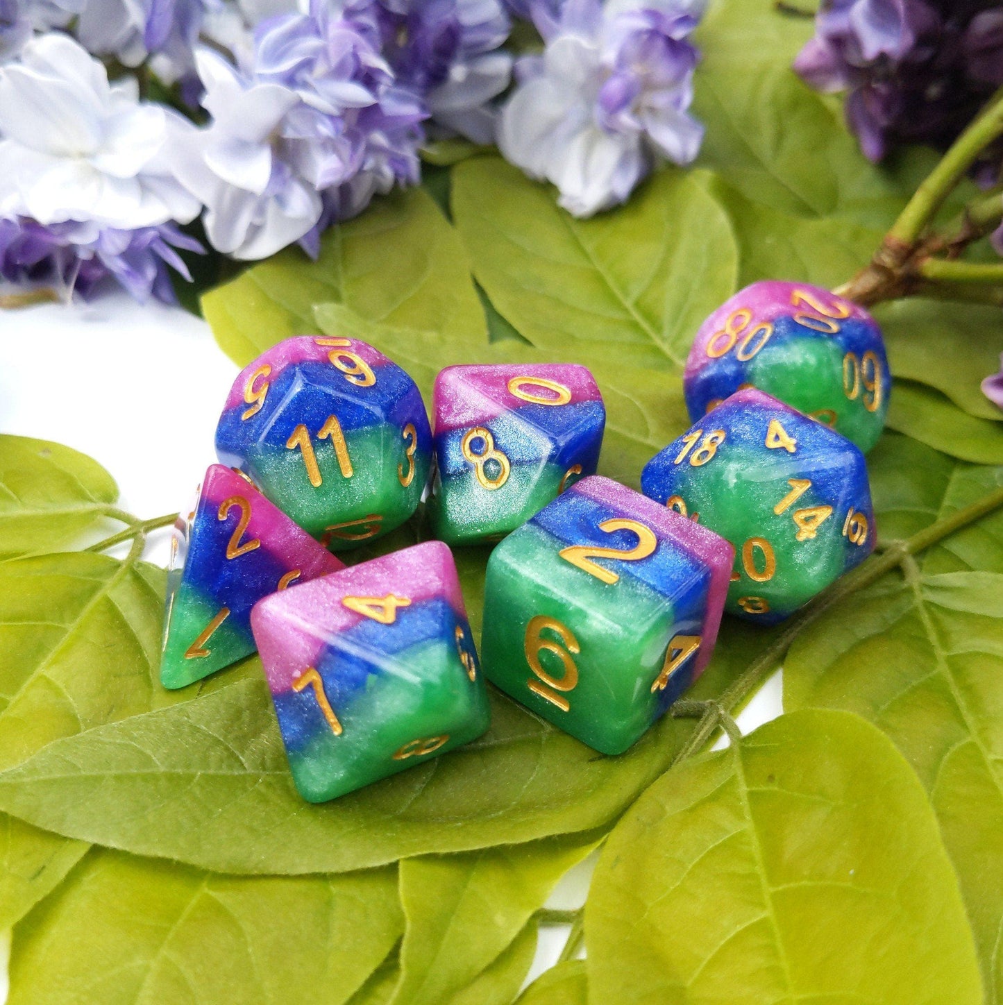 Jester's Gambit Dice Set in pearly pink, blue, and green with gold numbers, displayed on green leaves, ideal for TTRPGs.
