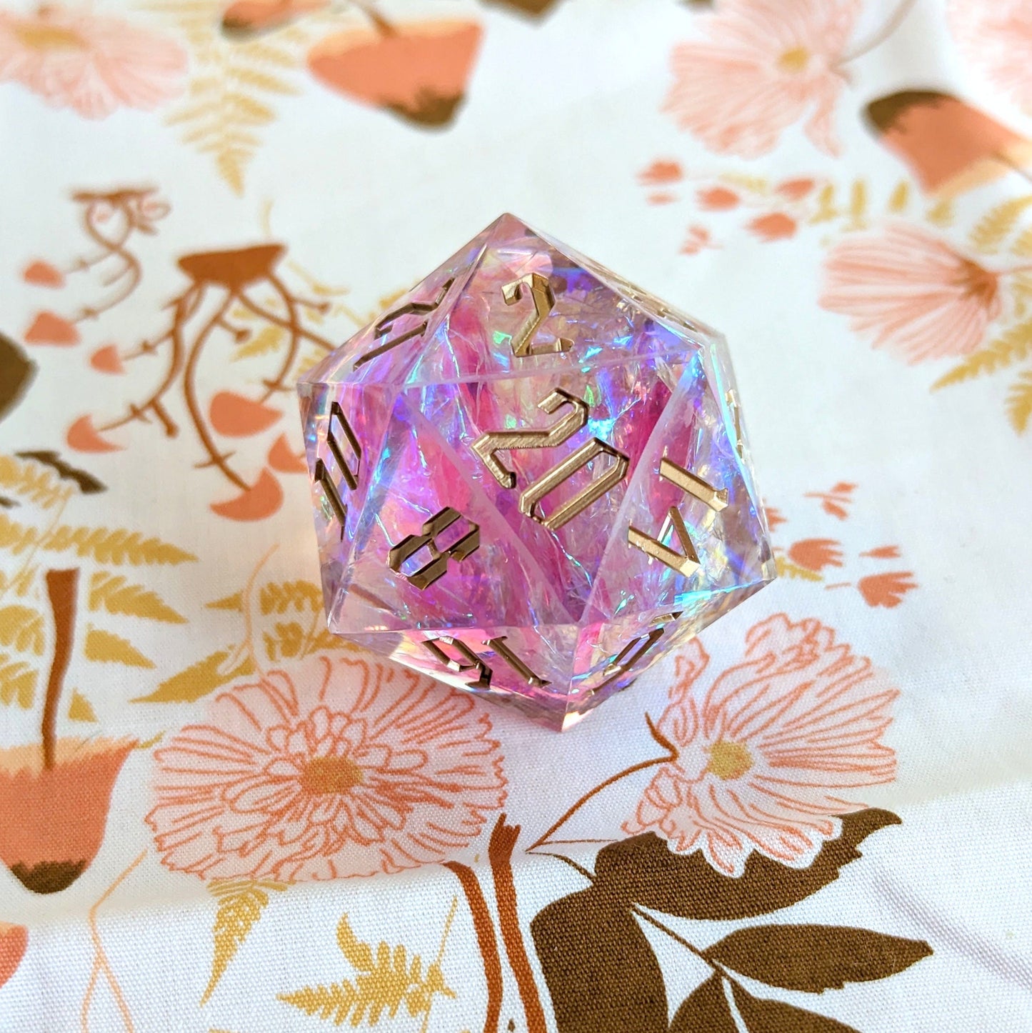 Large pink iridescent D20 sharp edge resin die with gold numbers for Dungeons and Dragons on floral background.