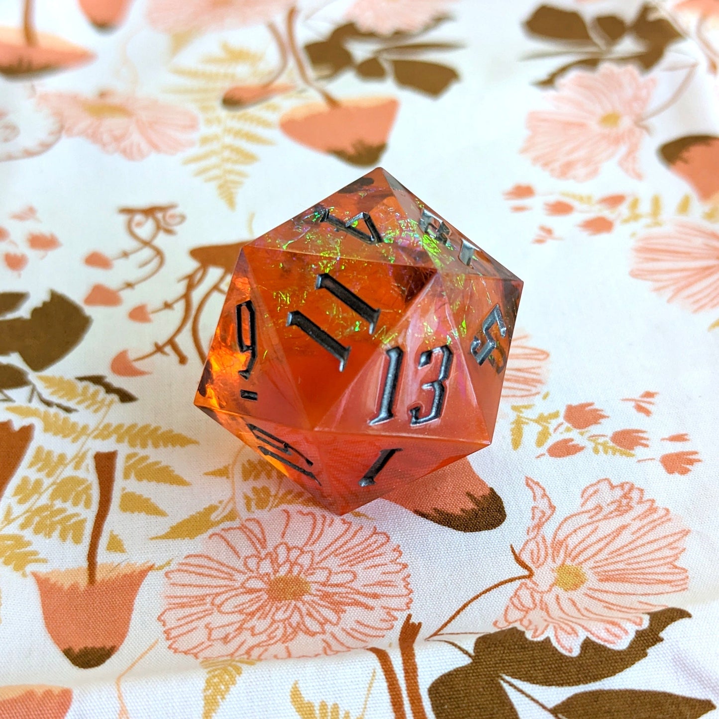 Orange sharp edge resin D20 die for Dungeons and Dragons, resting on a floral background, large 20-sided die for tabletop games.