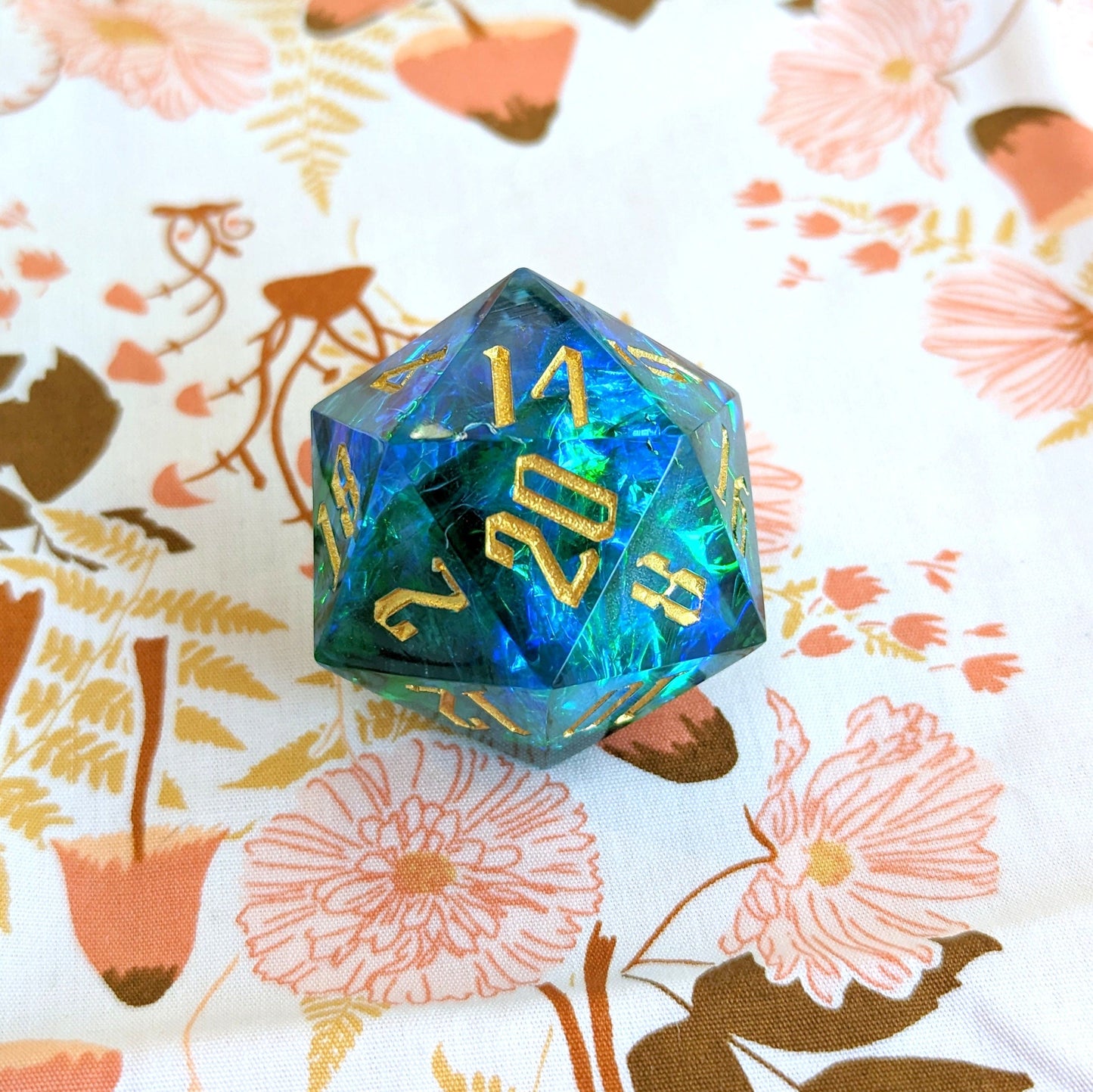 Large sharp edge blue and green D20 die for Dungeons and Dragons tabletop games on floral fabric background.