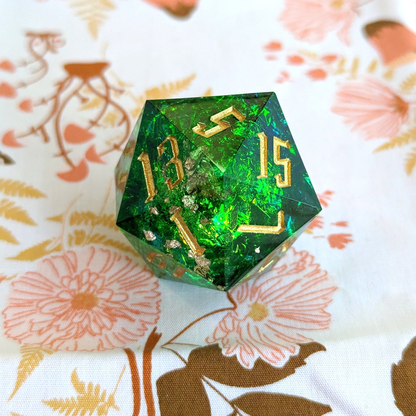 Large green sharp edge resin D20 die with gold numbers for Dungeons and Dragons on floral background.