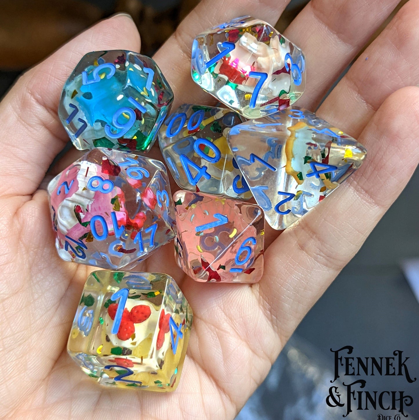 Hand holding Pastry Dice Set, 7 large resin dice with colorful miniature pastries inside, designed for tabletop RPGs like Dungeons & Dragons.