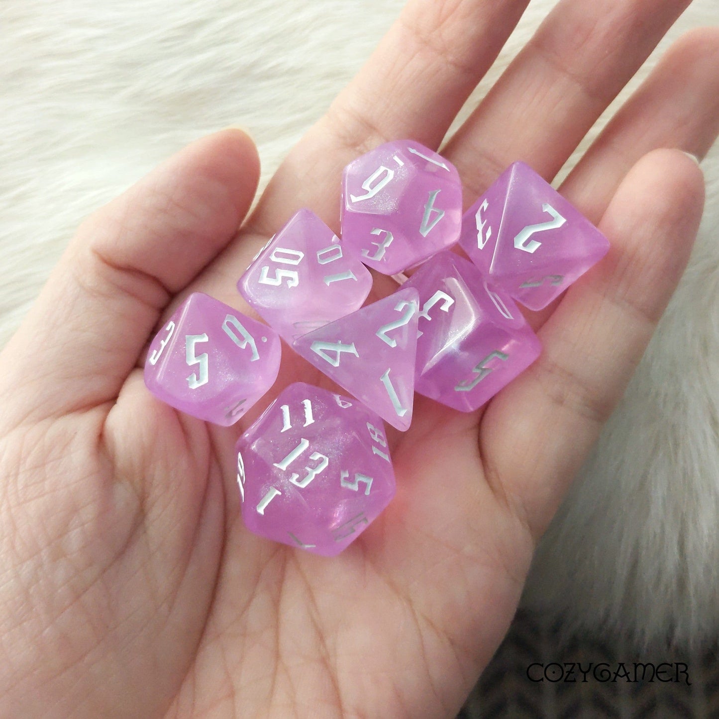 Hand holding pink glitter acrylic 7 piece polyhedral dice set with fantasy font for tabletop RPGs like Dungeons and Dragons.