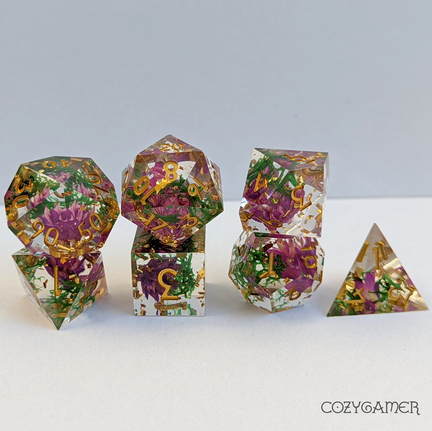 Purple flowers and green moss encased in clear resin sharp edge dice set, 7-piece polyhedral TTRPG dice with gold numbering.