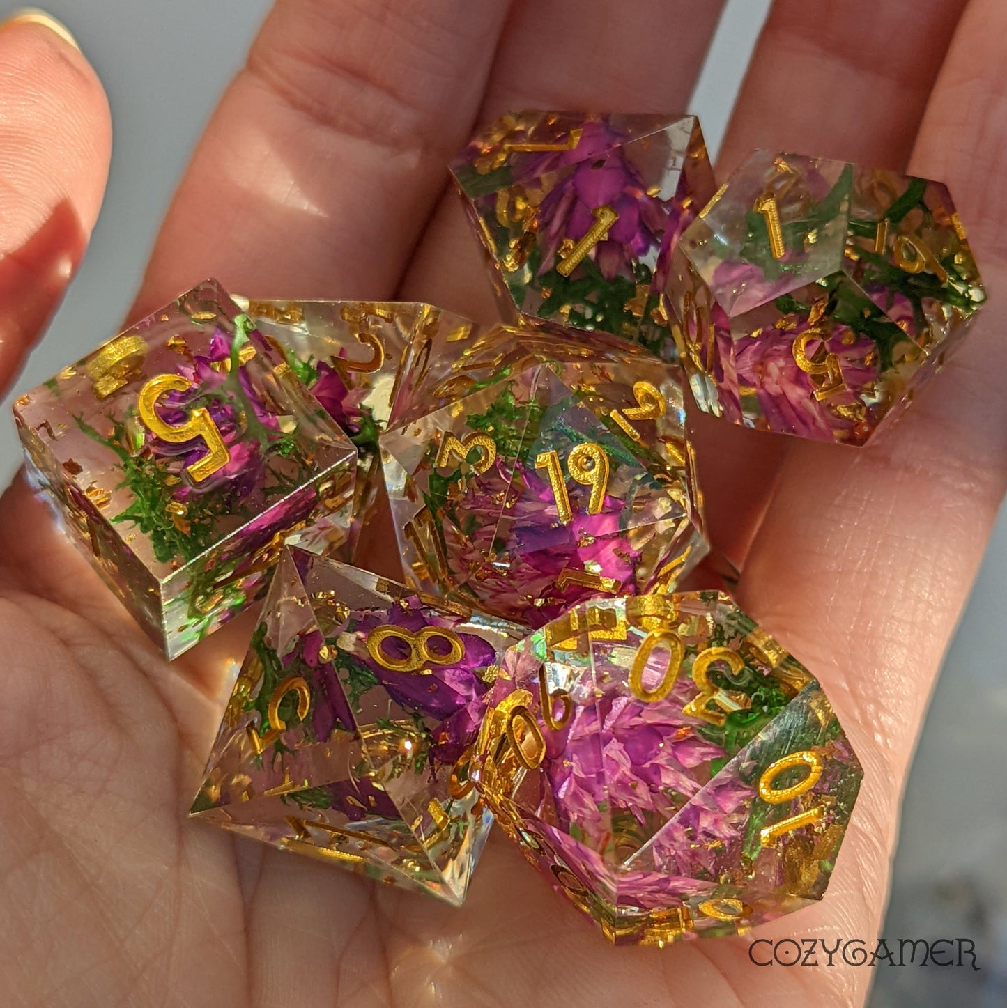 Hand holding sharp edge resin dice set with purple flowers and green moss. 7 piece DND polyhedral dice catching light.