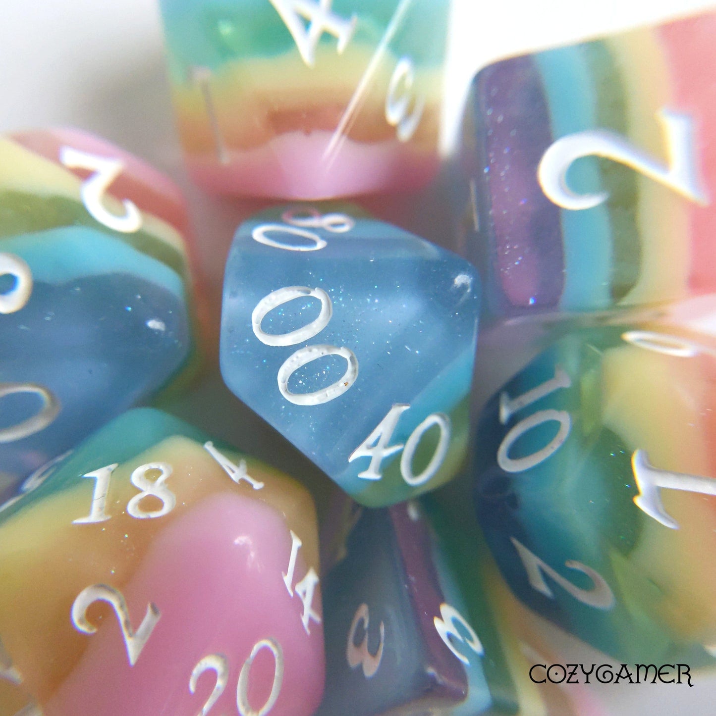 Pastel rainbow stripe DnD dice set with glittering layers and white numbers, featuring CozyGamer branding in the corner.