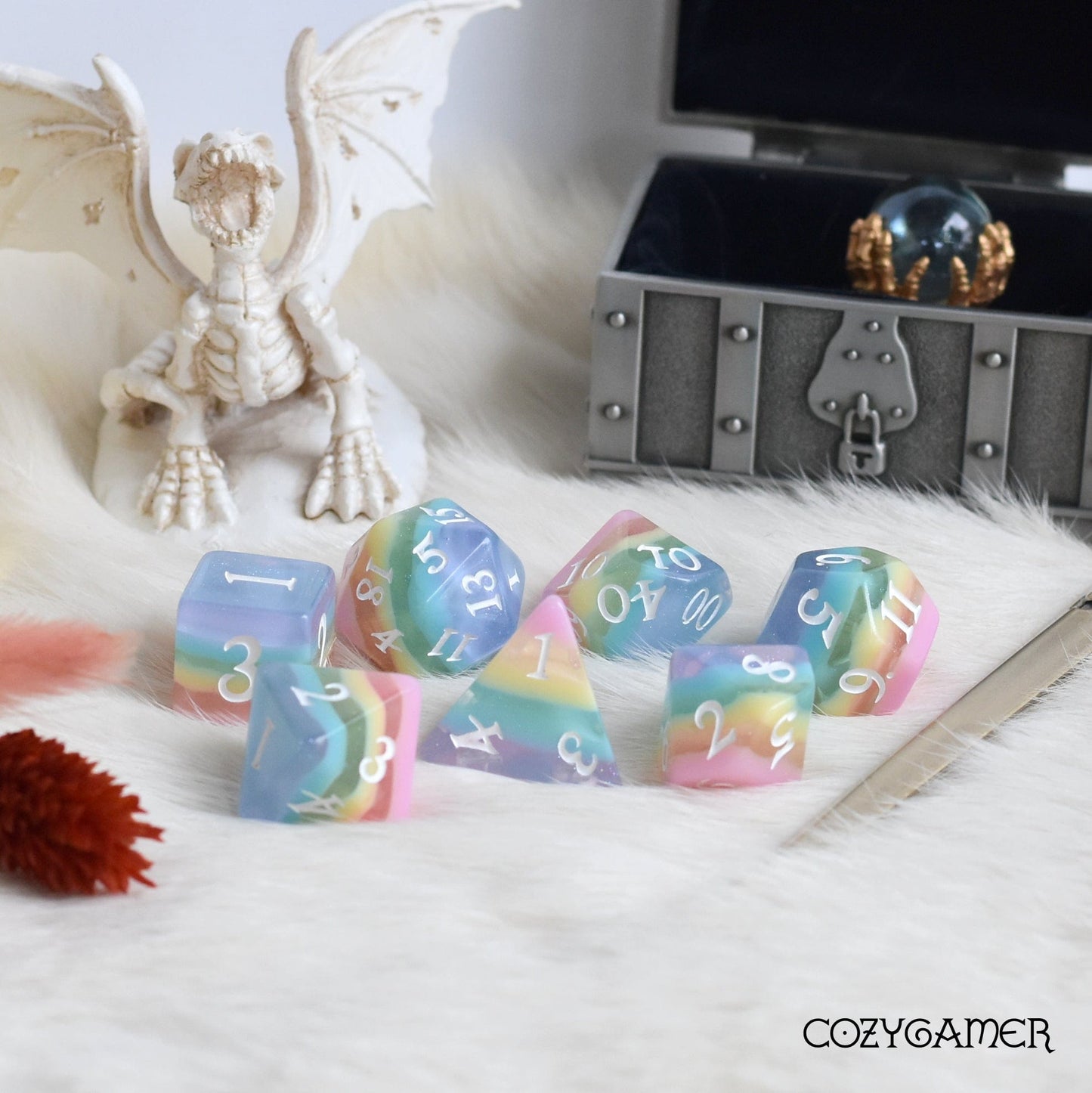 Pastel rainbow stripe DND dice set with glitter and a dragon figurine beside a treasure chest on a white fur surface.