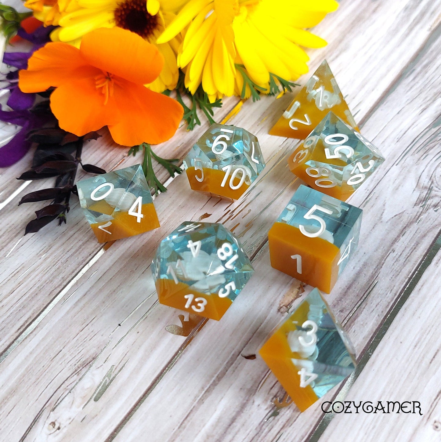 Sea Shell Sharp Edge Resin Dice Set with water and sand layers, 7 piece polyhedral DND TTRPG dice set displayed with colorful flowers.