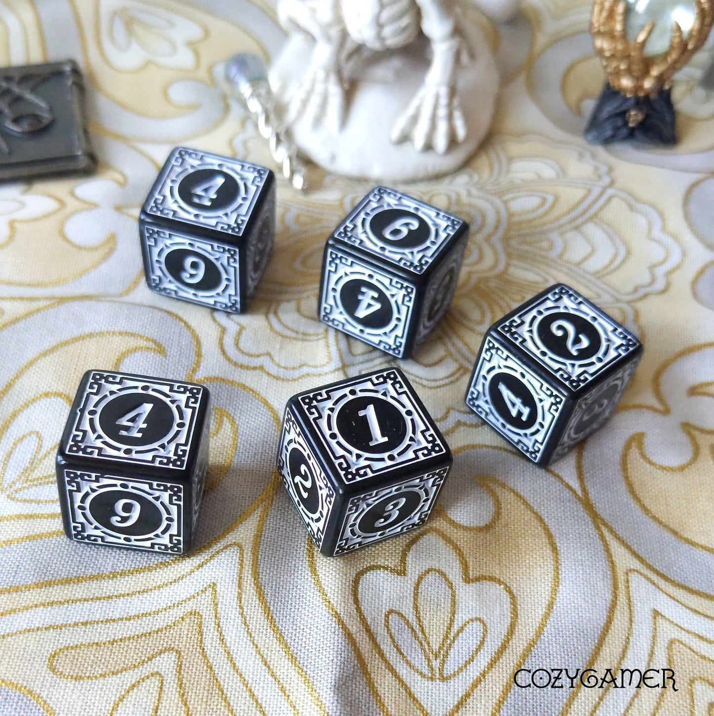Set of five black D6 dice with decorative frames on an ornate tablecloth.