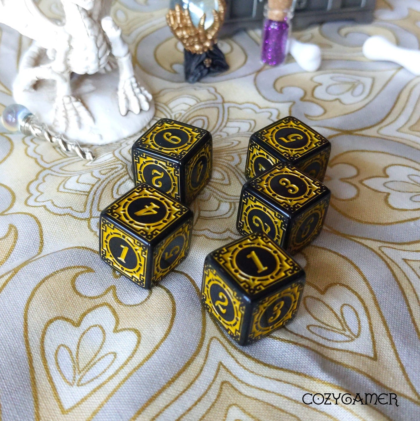 Black and gold ornate set of 5 six-sided dice on a patterned cloth background.