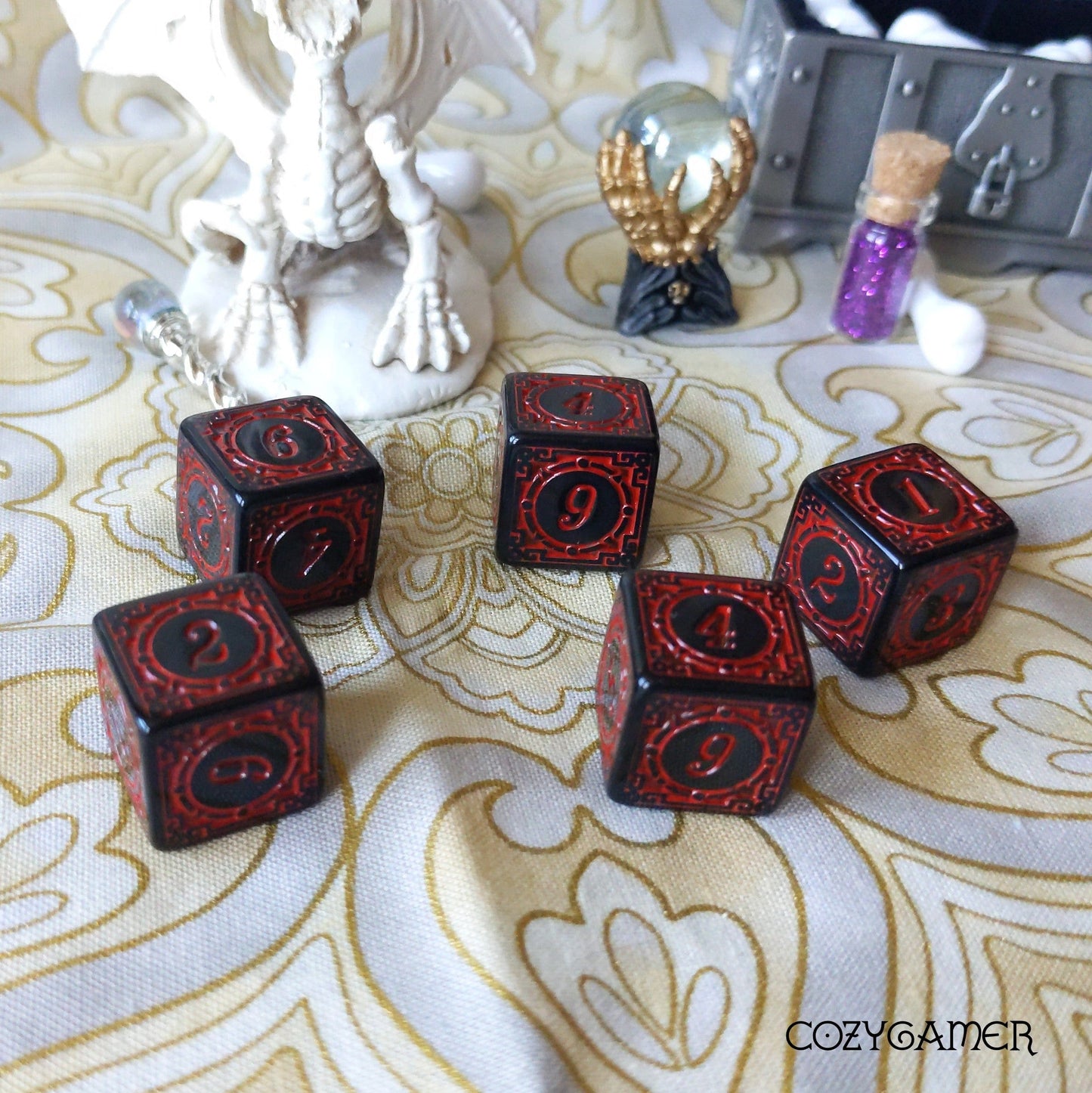 Black and red set of 5 D6 dice with ornate frames, displayed on patterned cloth with fantasy-themed decorations in the background.