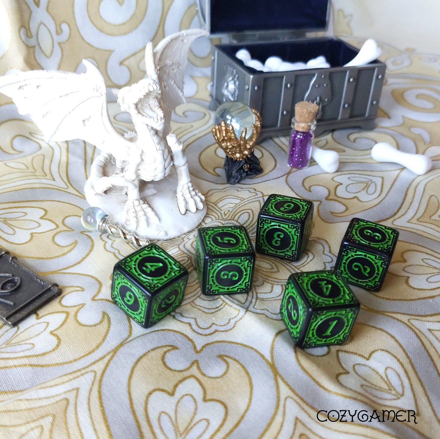 Green patterned D6 dice set with fantasy dragon figure and treasure chest backdrop on decorative table.